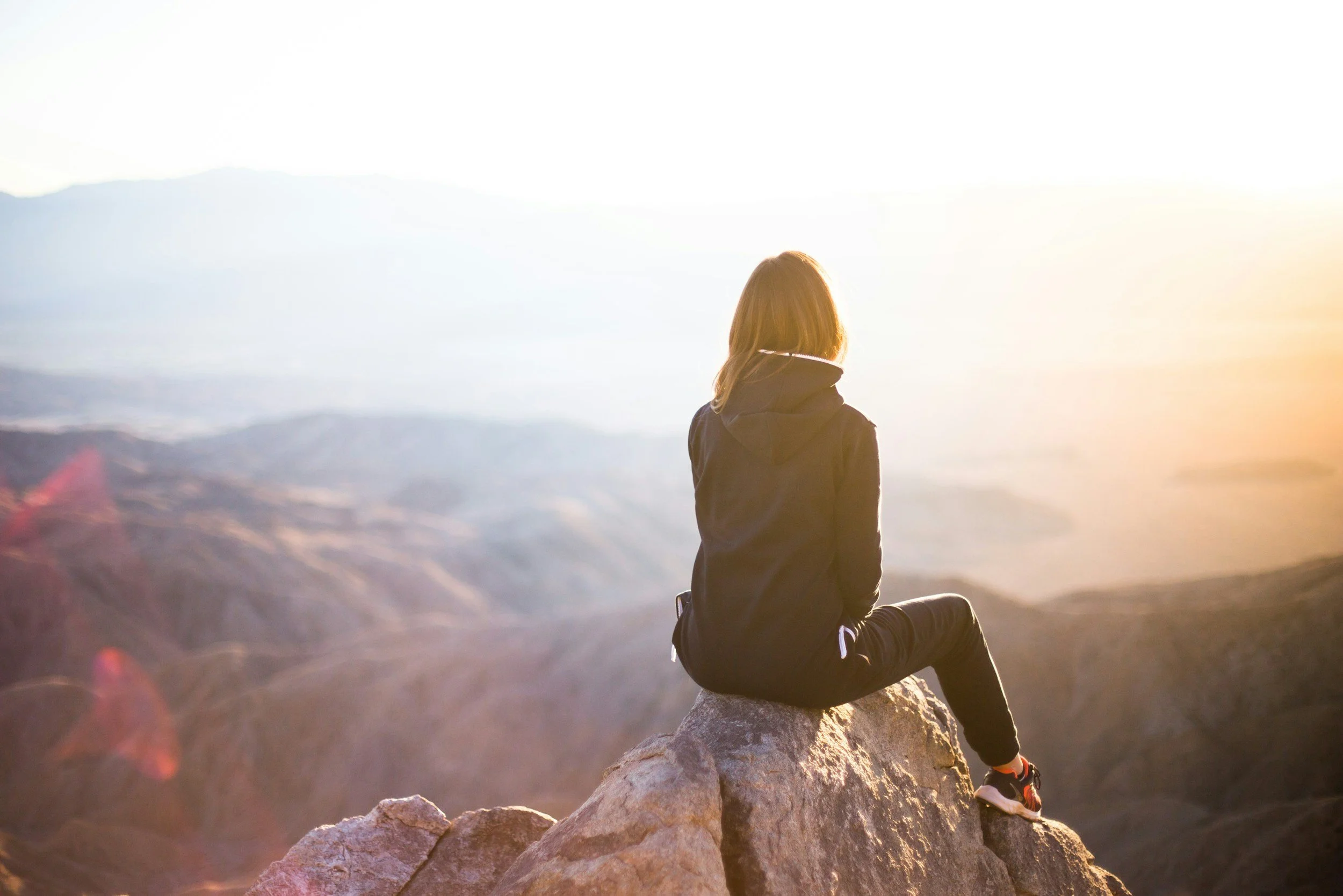 A woman sitting on a rock ledge overlooking a sunset or sunrise over a mountainous landscape.