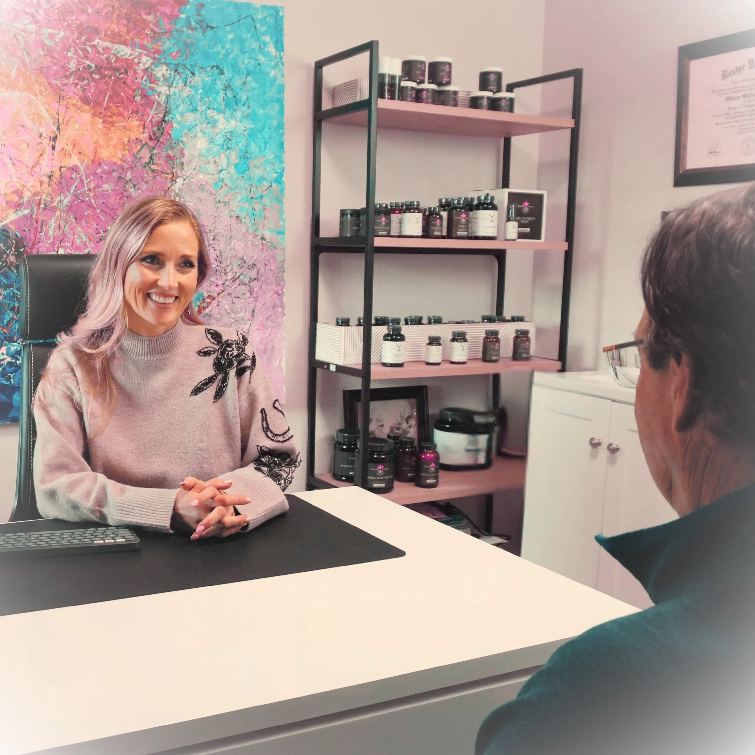 A woman with blonde hair smiling at a man wearing glasses during a consultation in an office with a colorful abstract painting on the wall and shelves with bottles of supplements or vitamins.