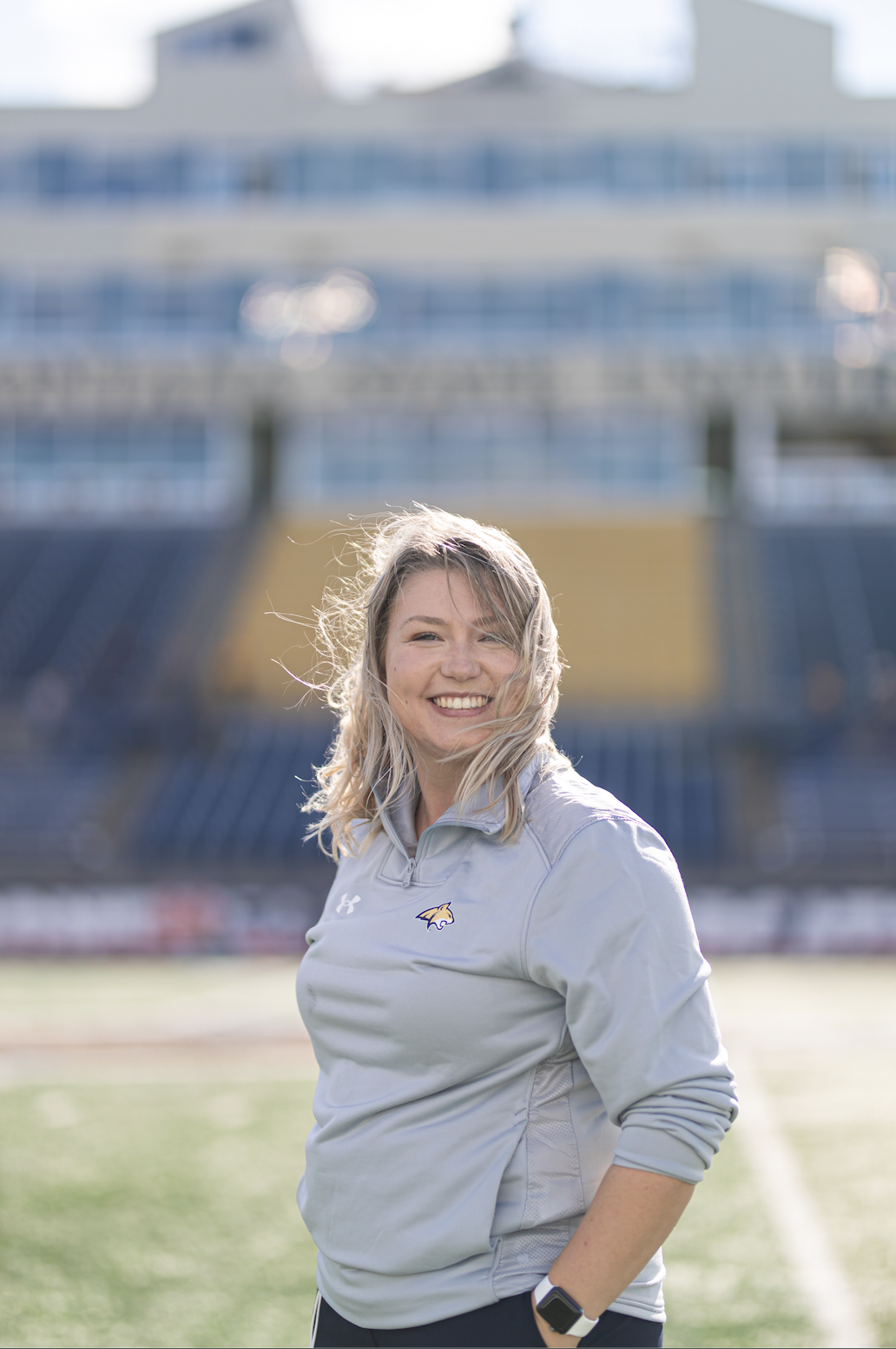 A young woman with blonde hair smiling on a sports field, wearing a light gray sports jacket with a logo, and a smartwatch on her wrist, with a stadium in the background.