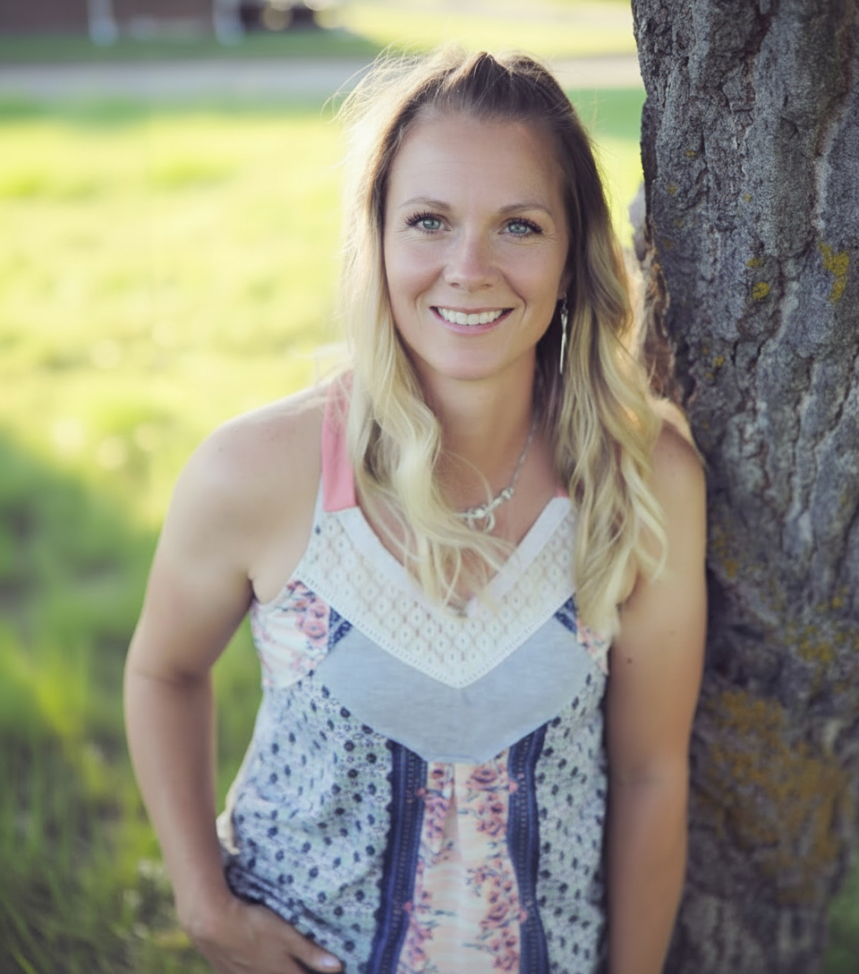 A woman with long blonde hair smiling, leaning against a tree outdoors in a park or garden.
