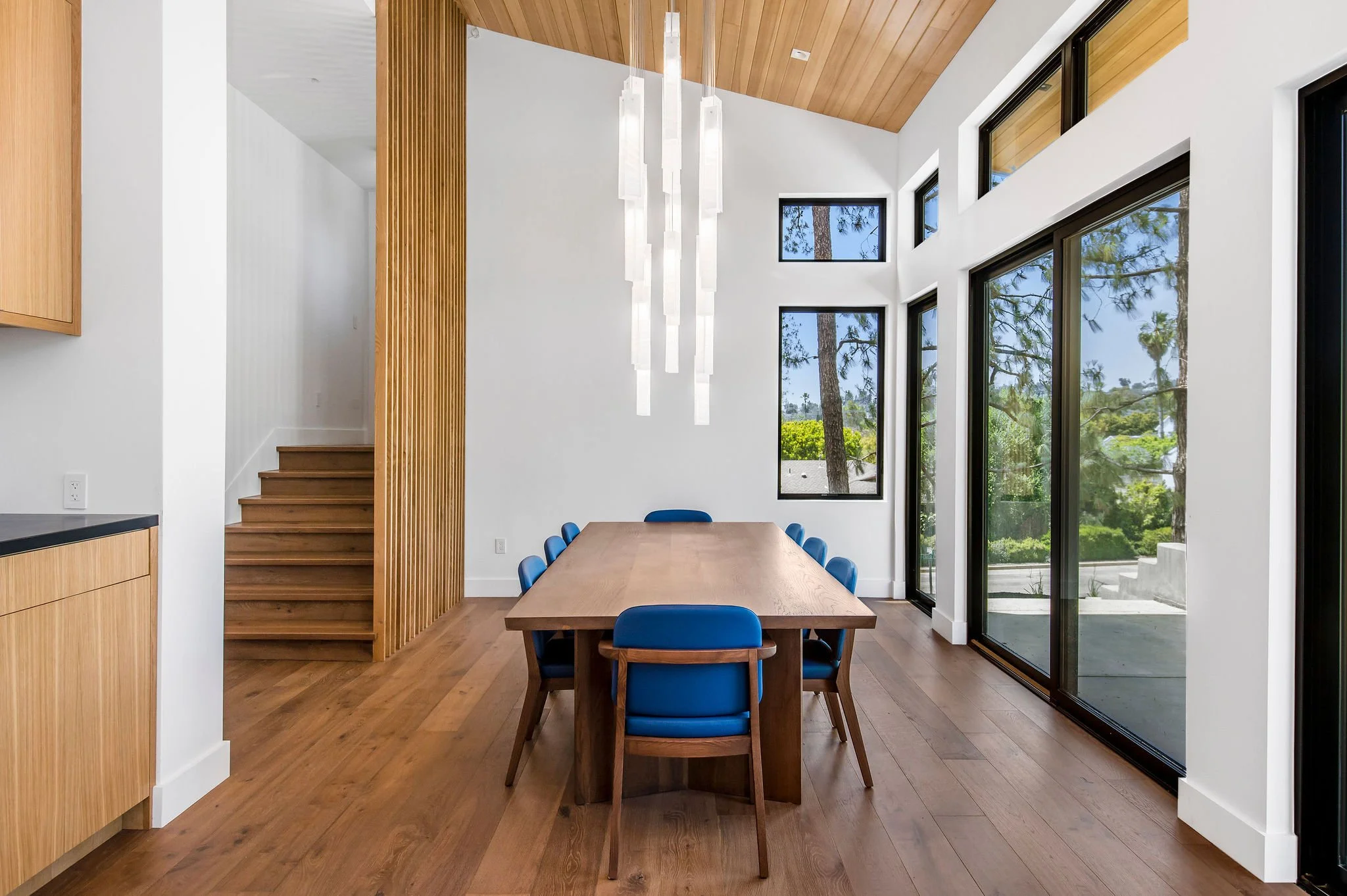 Modern dining room with a long wooden table, blue chairs, sleek chandelier, and large windows overlooking trees.
