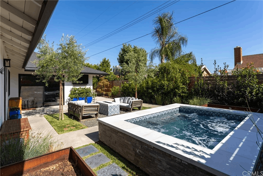 Backyard with a small, elevated pool, outdoor seating, and green landscaping under a clear blue sky.