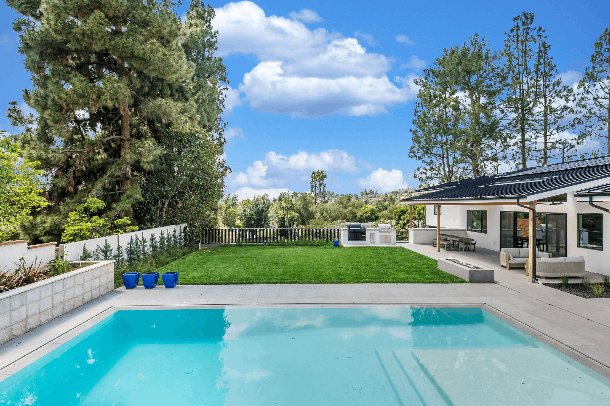 Modern backyard with a sparkling blue pool, green lawn, outdoor kitchen, patio, and a white house under a blue sky.