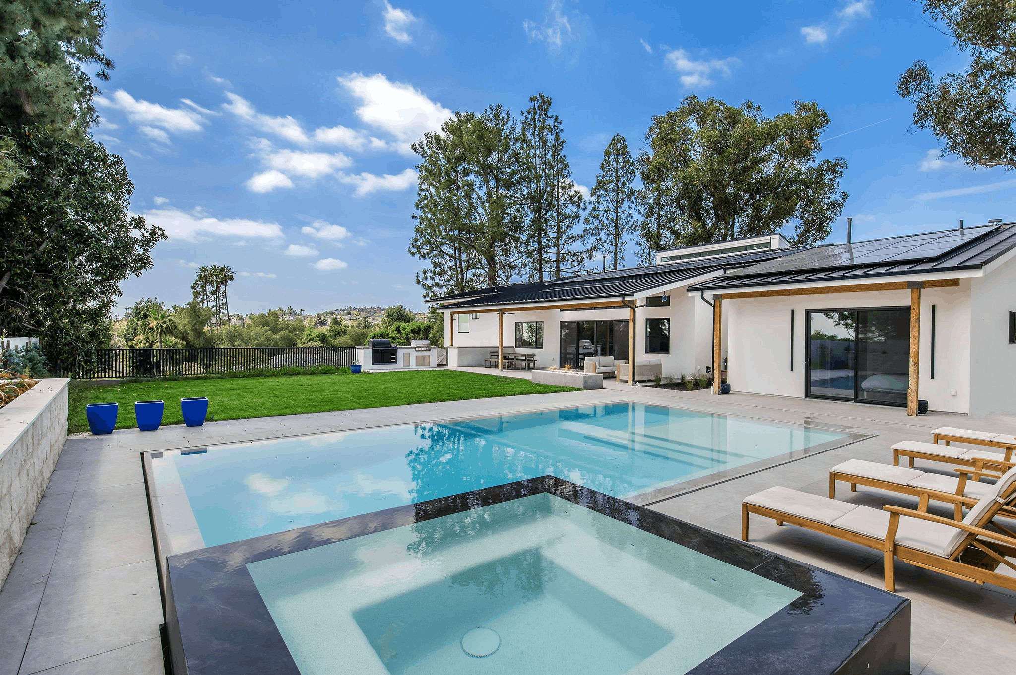 Modern backyard with swimming pool, spa, lounge chairs, green lawn, outdoor kitchen, and white house with solar panels.