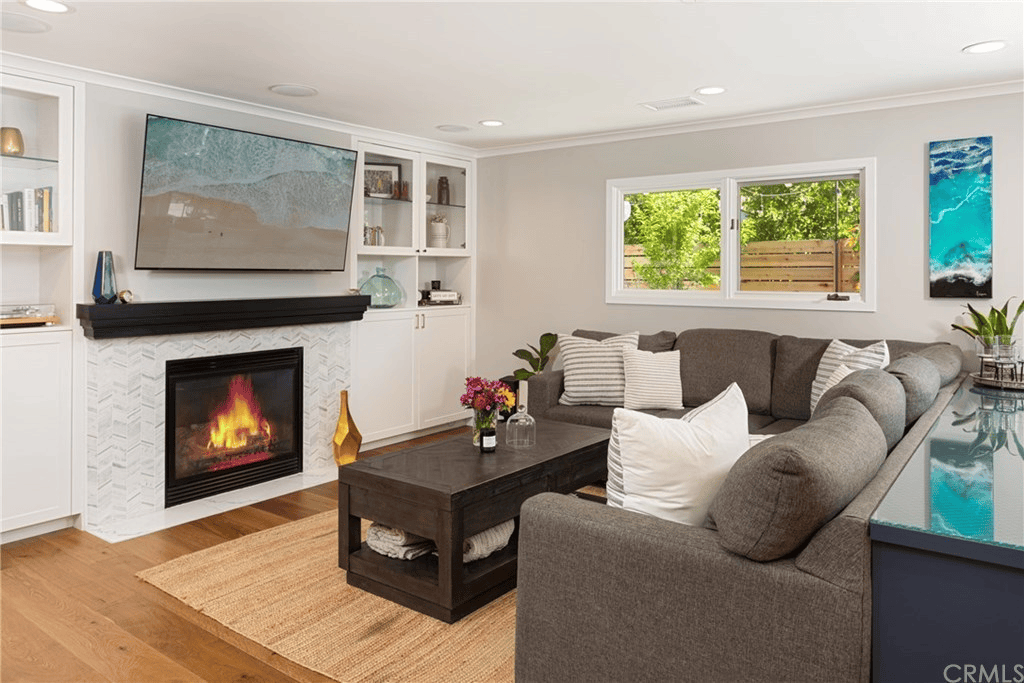 A cozy living room with a fireplace, TV displaying a beach scene, a large sectional sofa, and a wooden coffee table.
