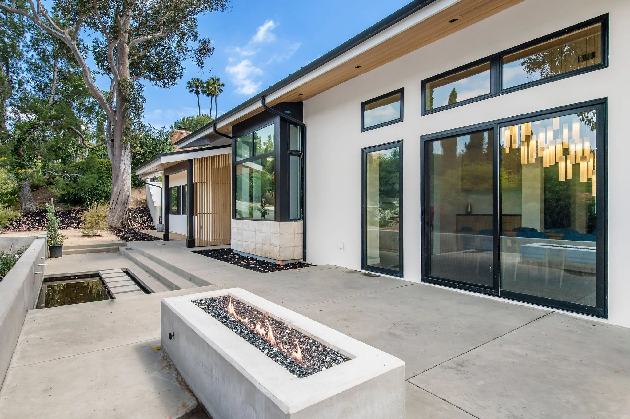 Modern house with white walls, black-framed windows, and a concrete patio with a fire pit and water feature.