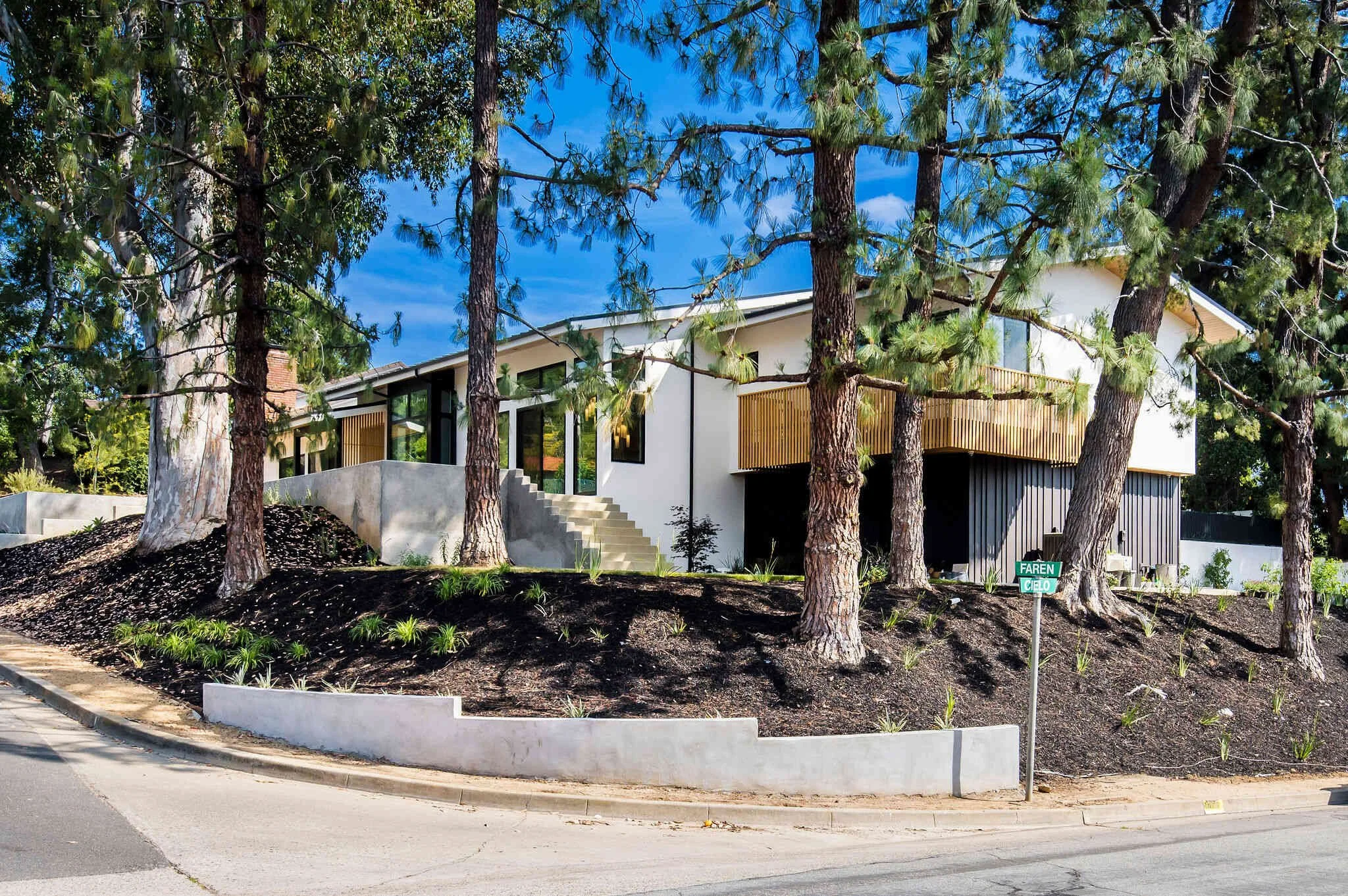 Modern white house with large windows and a wooden-slatted balcony, surrounded by pine trees and dark mulch.