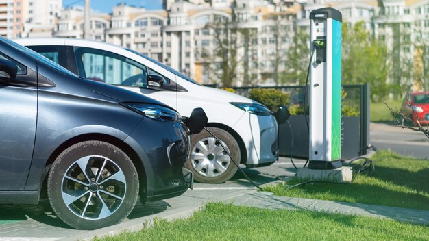 Two electric cars, one white and one dark grey, charging at a public station with buildings in the background.