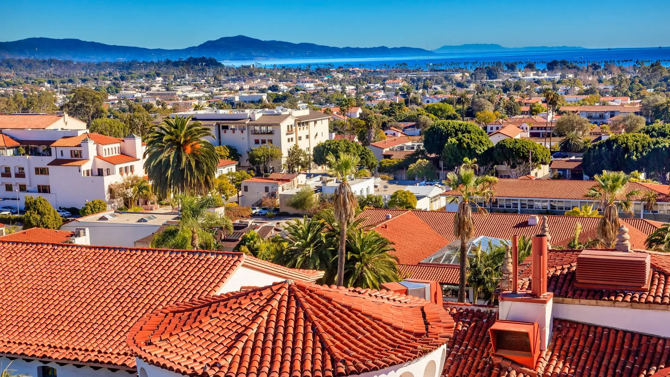 Aerial view of Santa Barbara: red tile roofs, palm trees, mountains, and the Pacific Ocean under a blue sky.