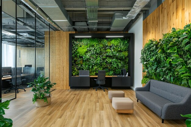 Modern office with wooden floors, glass walls, and two lush green living walls. A desk and a grey couch are visible.