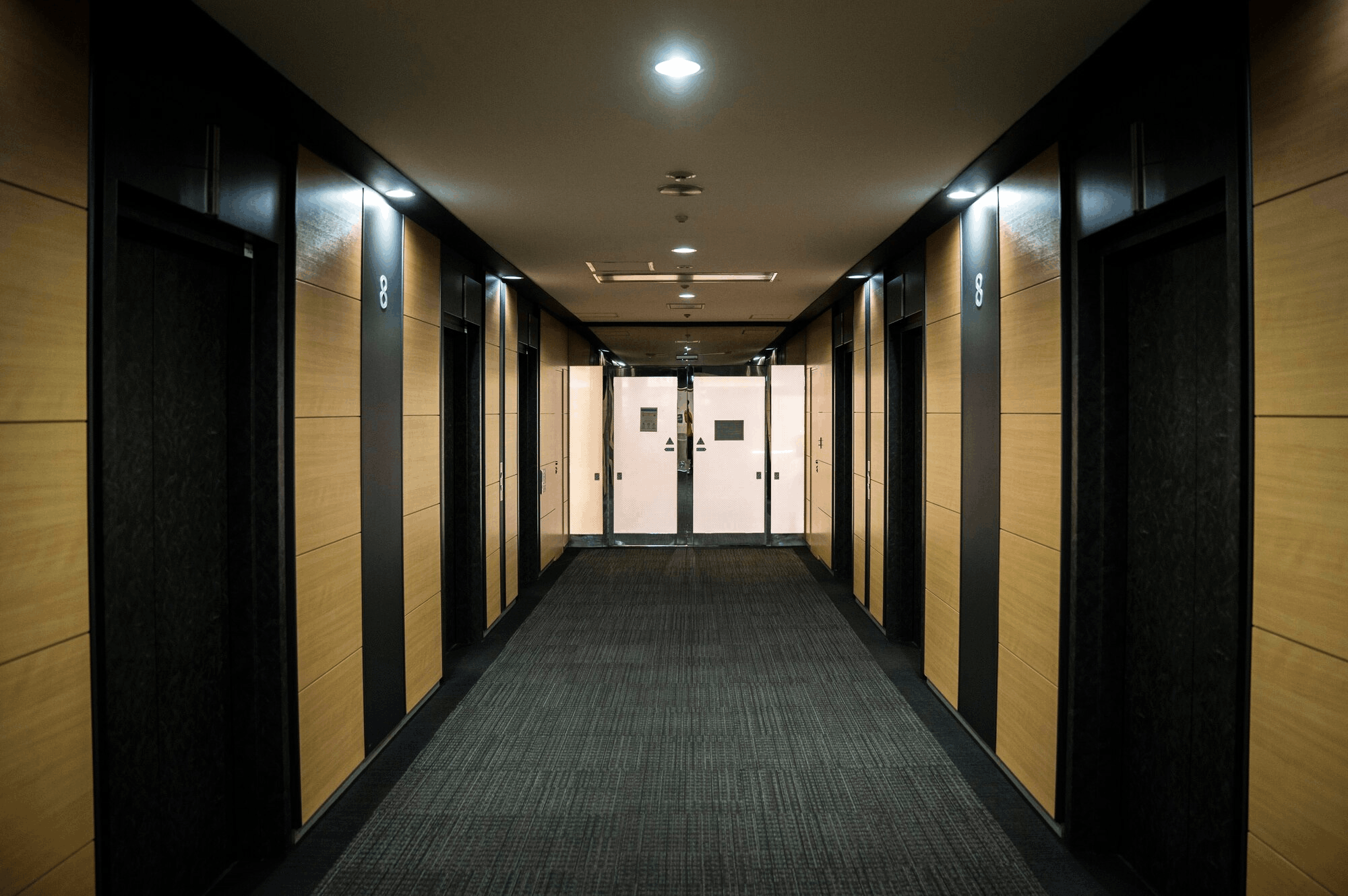 Long, empty office hallway with black doors and light wood paneling, leading to white double doors.