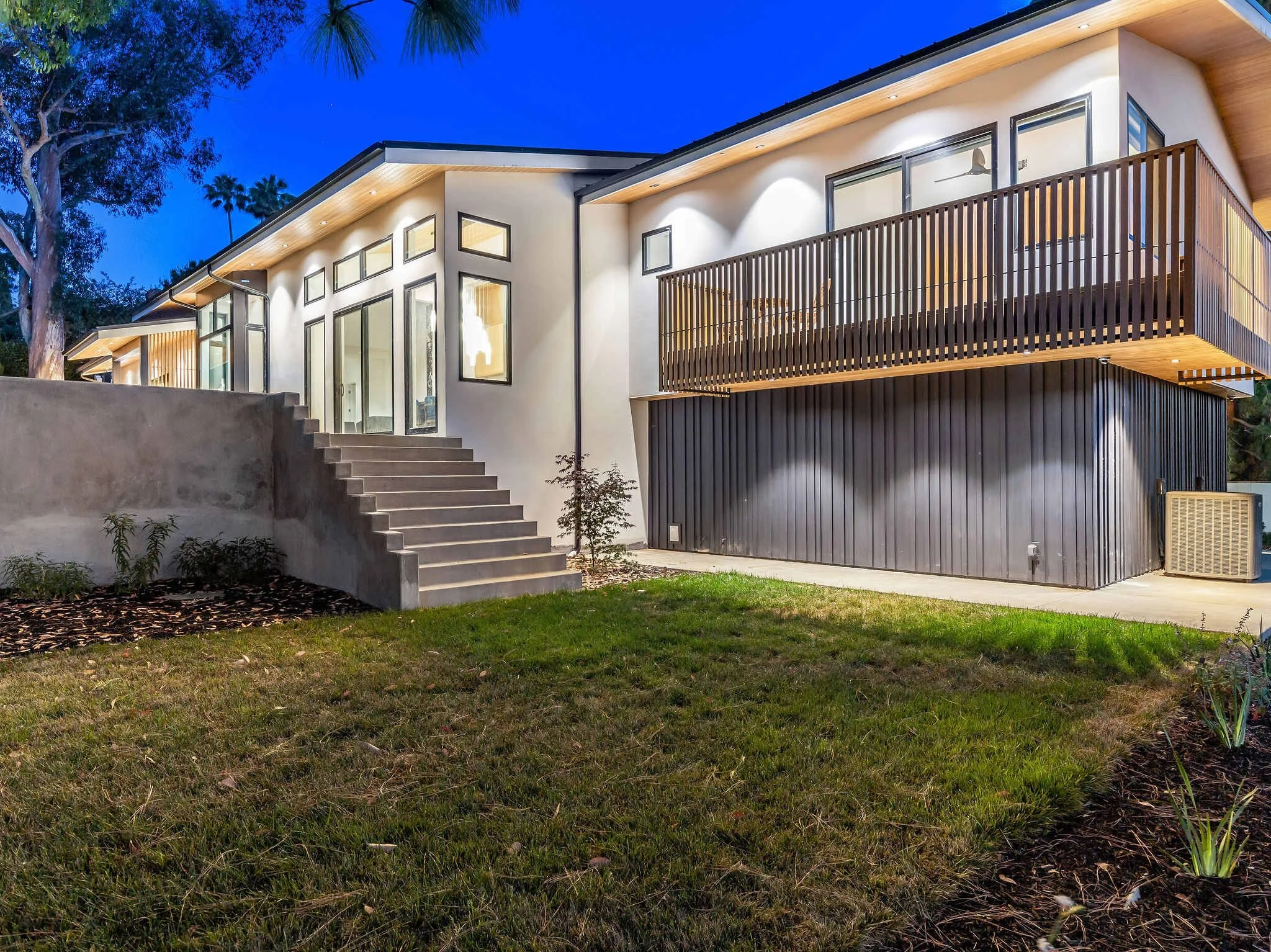 Modern house at dusk with concrete stairs leading to glass doors, a wood balcony, and a grassy yard.