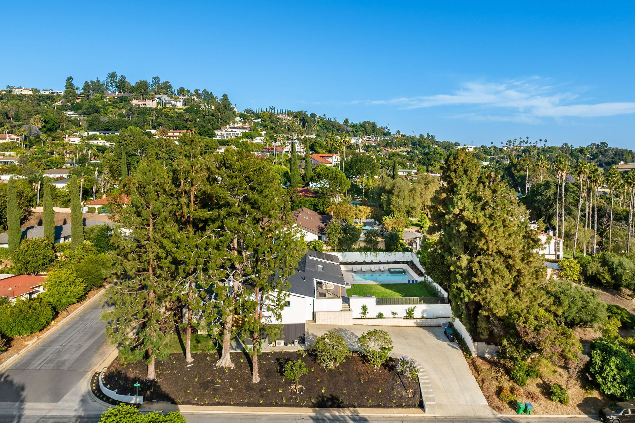 Aerial view of a white modern house with a pool and green lawn, surrounded by lush trees and houses on a hill.