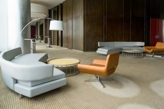 Modern office sitting area with light gray, curved sofas, orange chairs, round tables, and a large arc lamp on a patterned carpet.