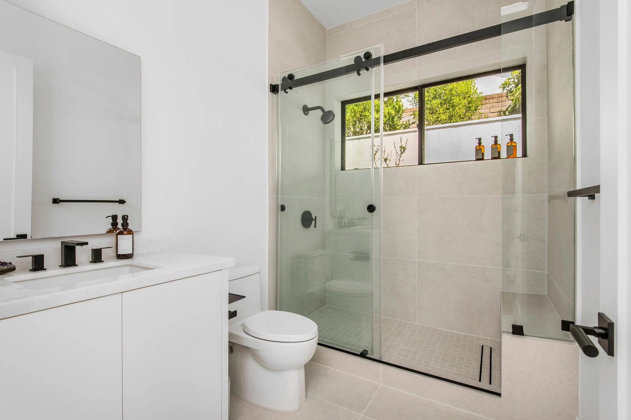Modern bathroom with white vanity, black fixtures, glass shower, and a window overlooking lush greenery.