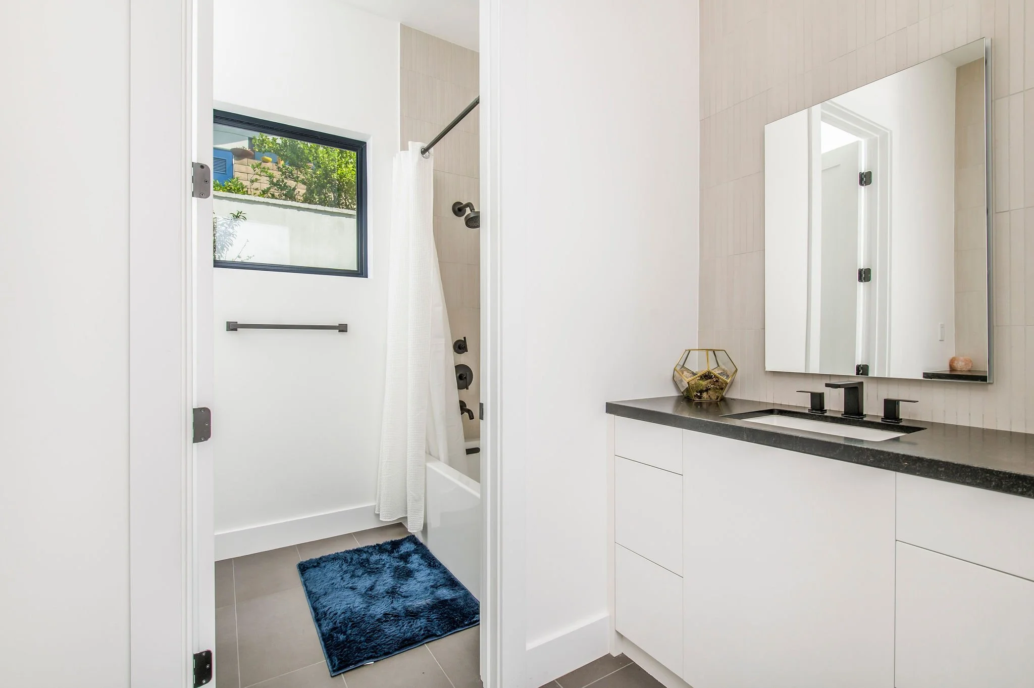 Modern white bathroom with black fixtures, a shower with a blue mat, and a vanity with a terrarium.