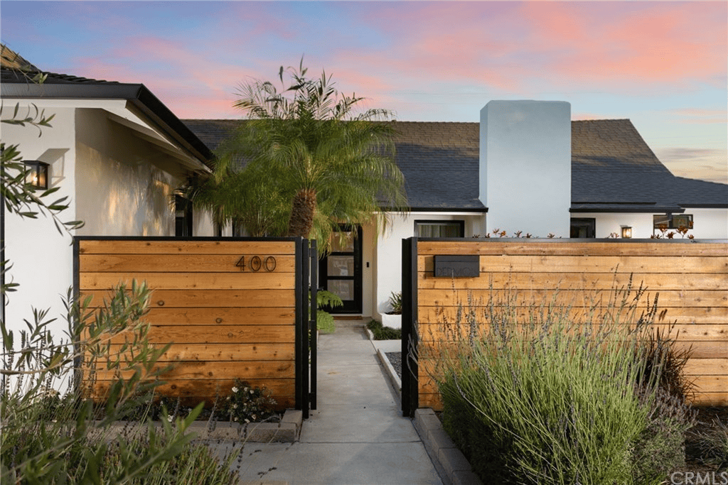 Modern white house with a dark roof behind a wooden fence with an open gate and a palm tree in the yard.