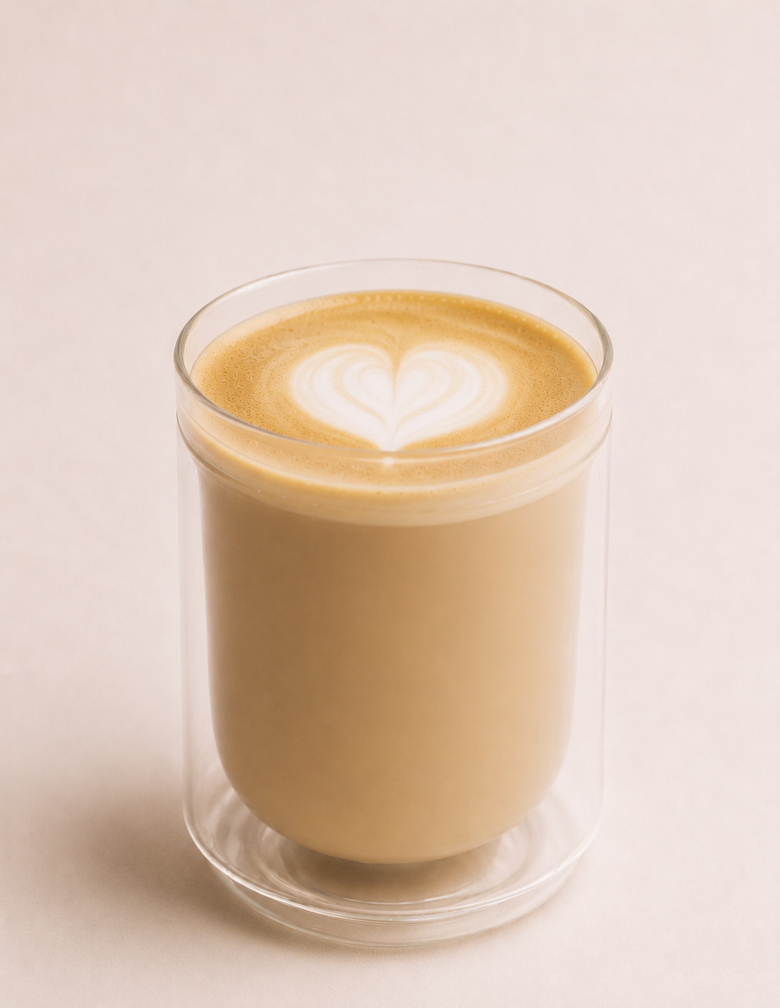 A clear glass of coffee with latte art in the shape of a heart, set against a plain light background.