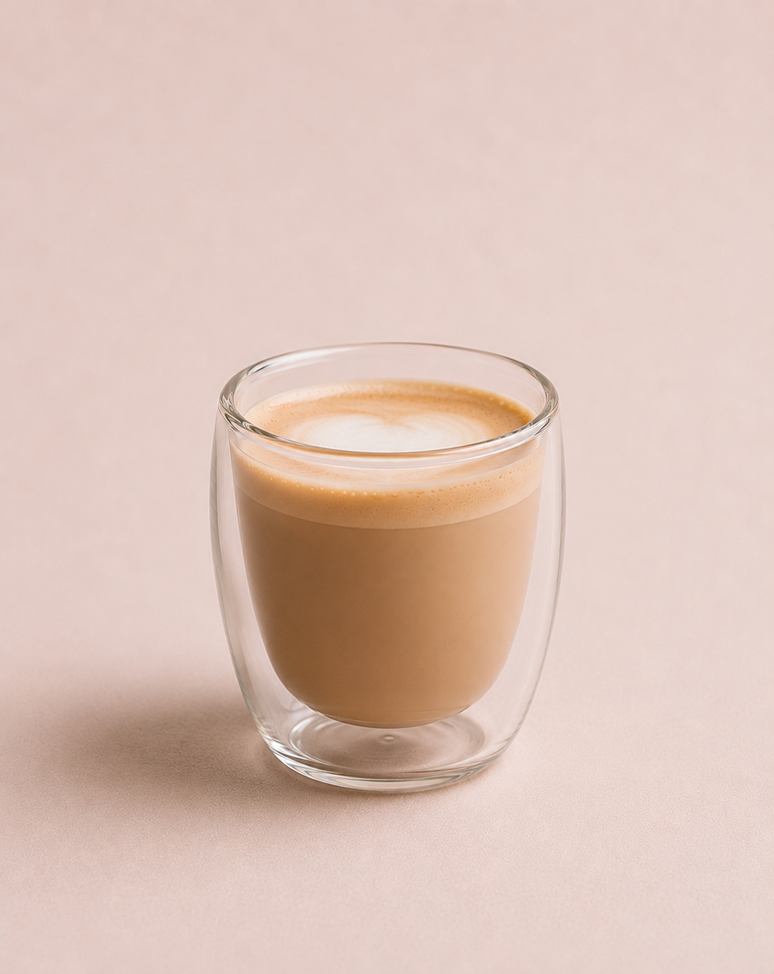 A clear glass cup filled with a frothy, light brown beverage, possibly coffee or tea, on a pale pink background.