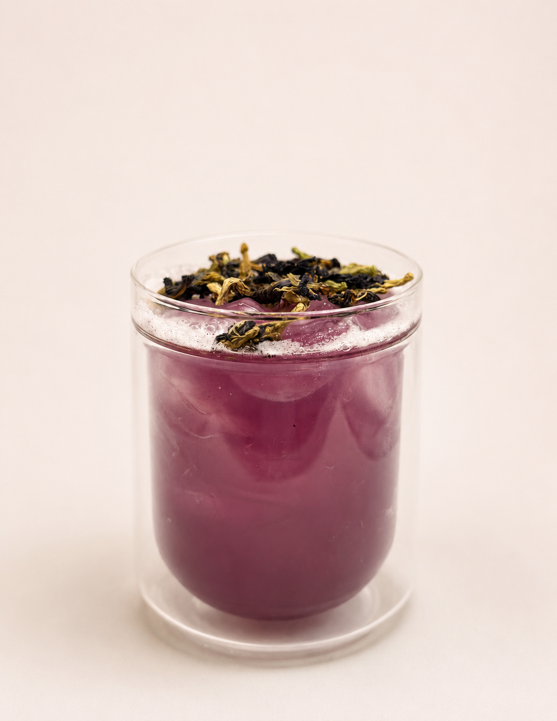 A clear glass filled with a purple or lavender-colored beverage, topped with dried and fresh purple flowers.