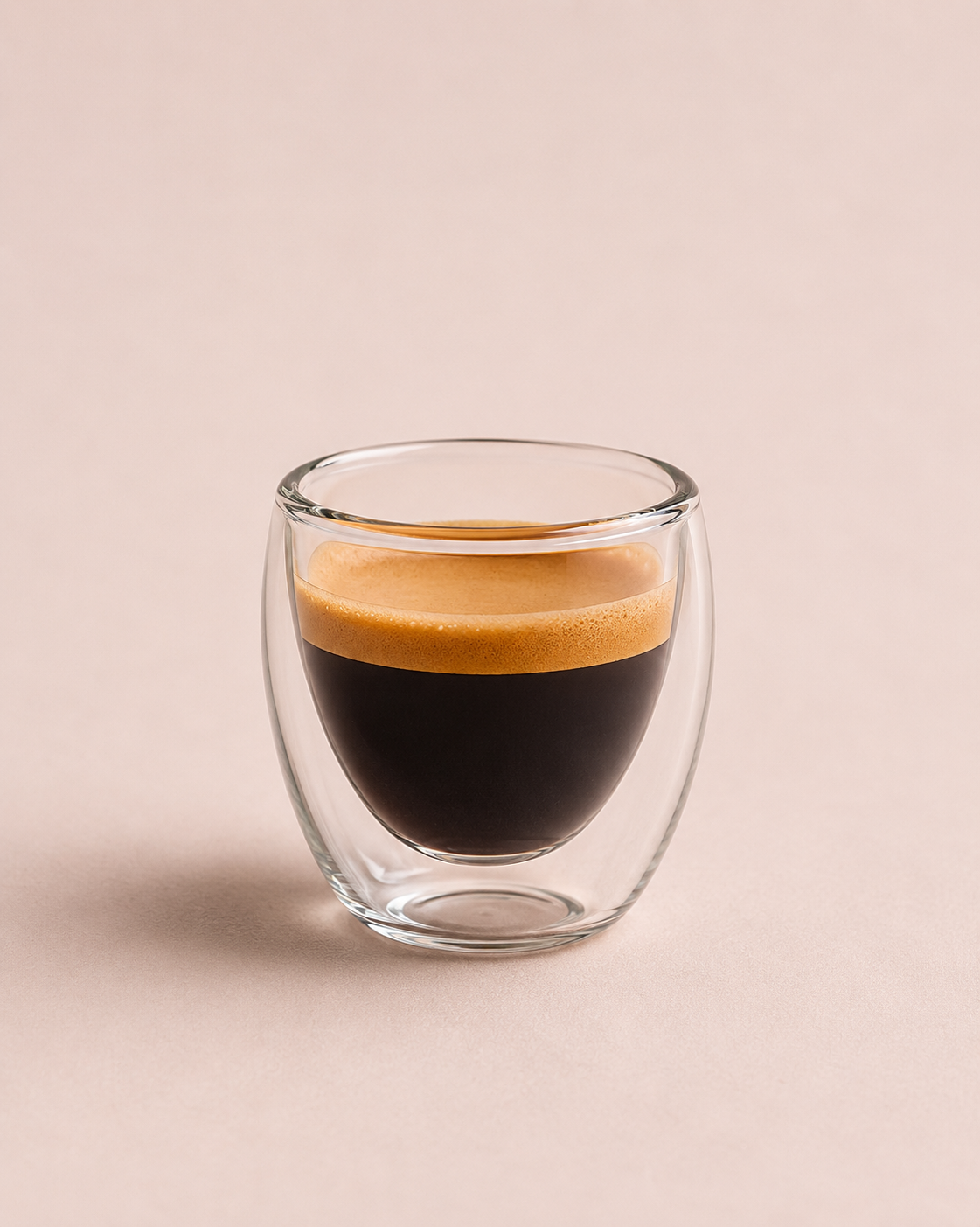 A clear glass cup with a dark espresso shot topped with a layer of crema, placed on a light pink surface.
