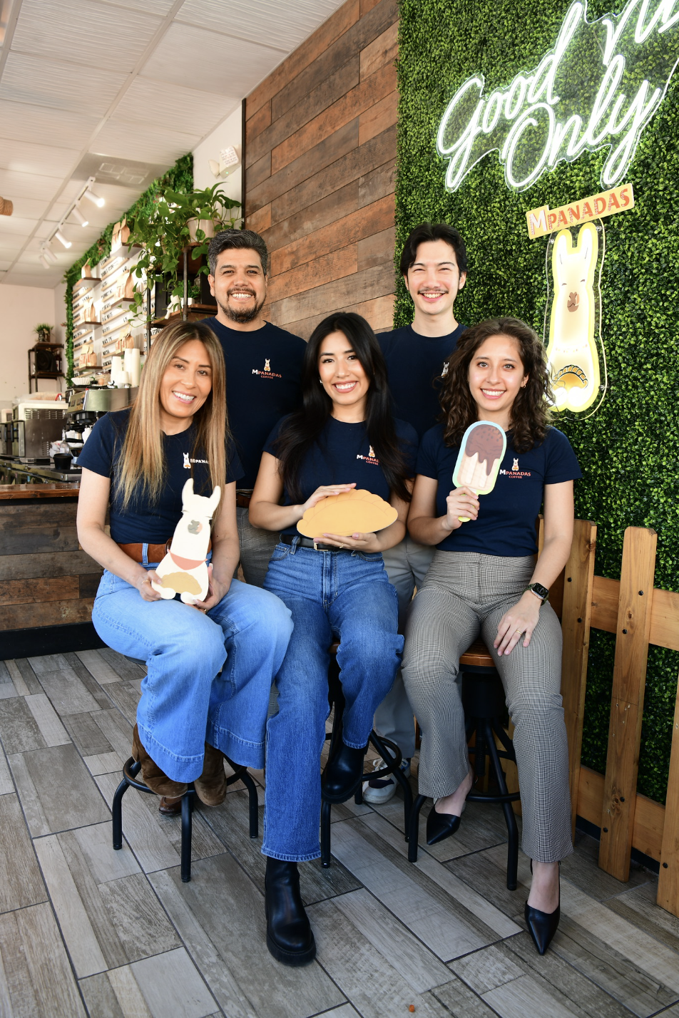 Group of five people smiling in a cafe, holding cartoon food and ice cream props, with a green wall and neon signs in the background.
