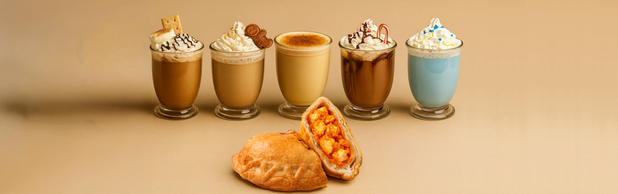 Six glasses of different topped coffee drinks arranged behind a wooden plate with a pumpkin pie and an empanada on it, on a cream-colored surface with shadow.