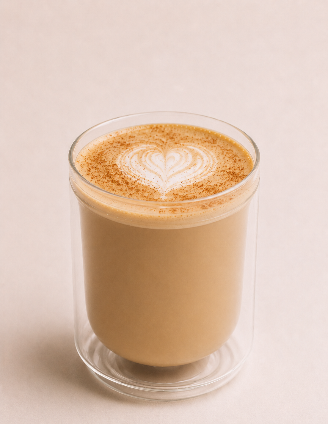 A clear glass filled with a creamy coffee beverage with latte art and sprinkled cinnamon on top, placed on a light background.