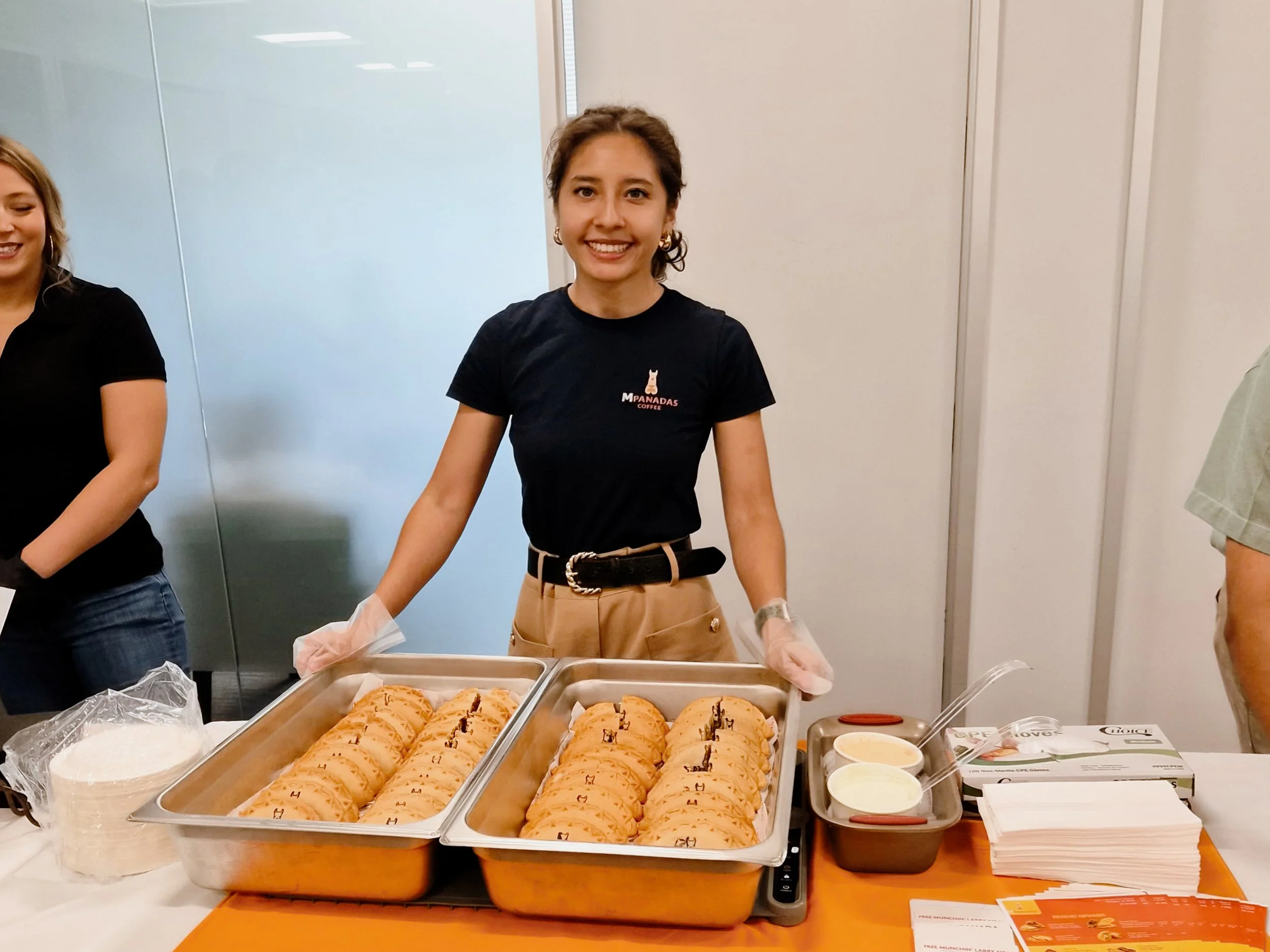 A woman smiling at a food stall with trays of cookies, cups of cream, and other items, wearing a black T-shirt with a logo that says 'MPANADAS COFFEE'.