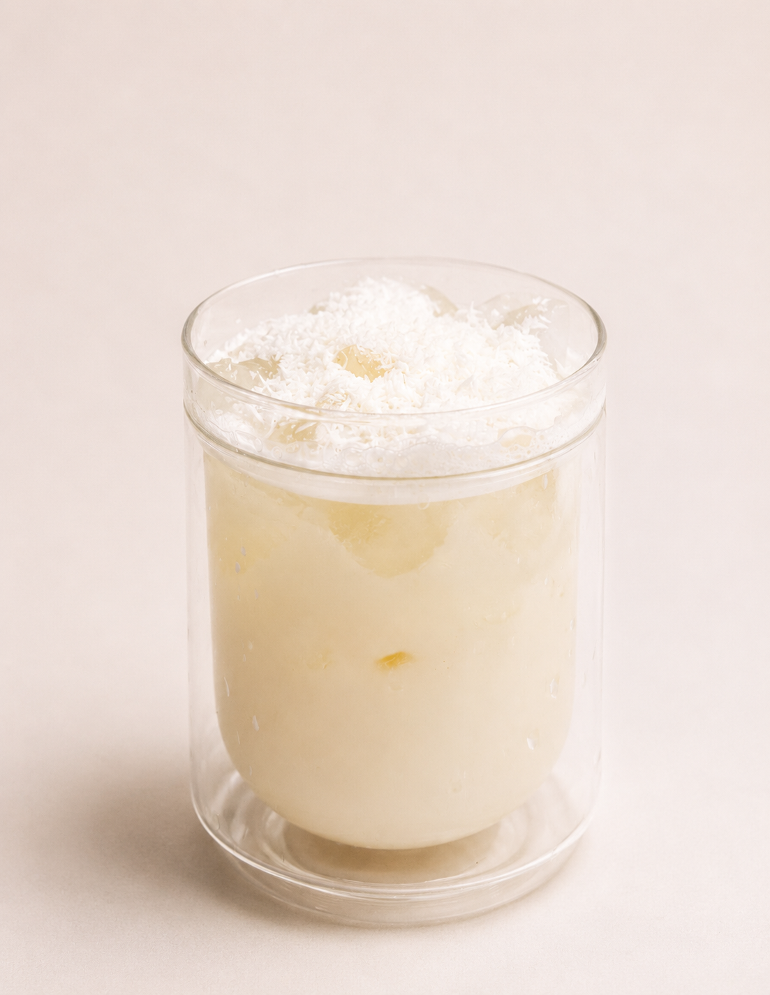 A clear glass of a creamy, pale yellow beverage topped with shredded white coconut.