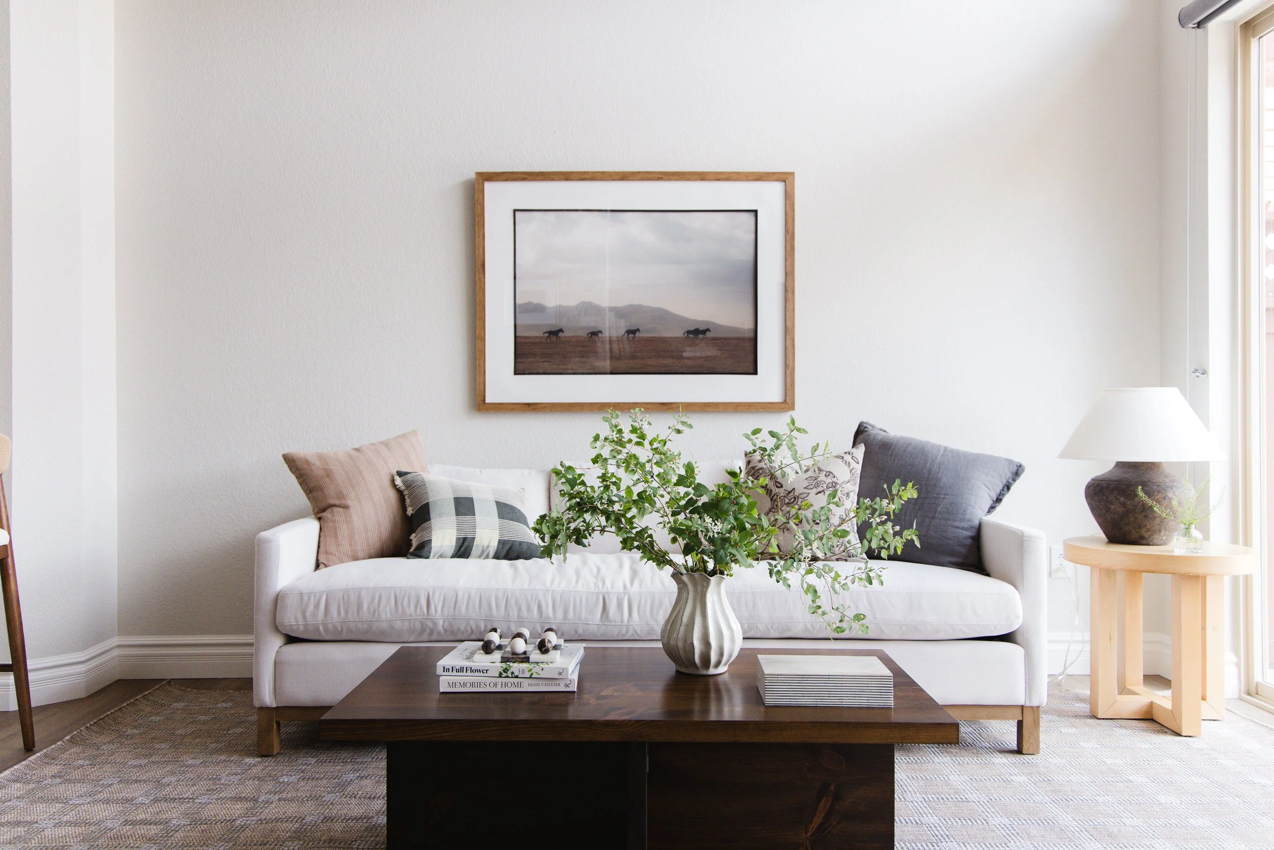Cozy Transitional Living room with western art, a down linen sofa, and rustic table lamp. There is a walnut coffee table with books and a vase with greenery.