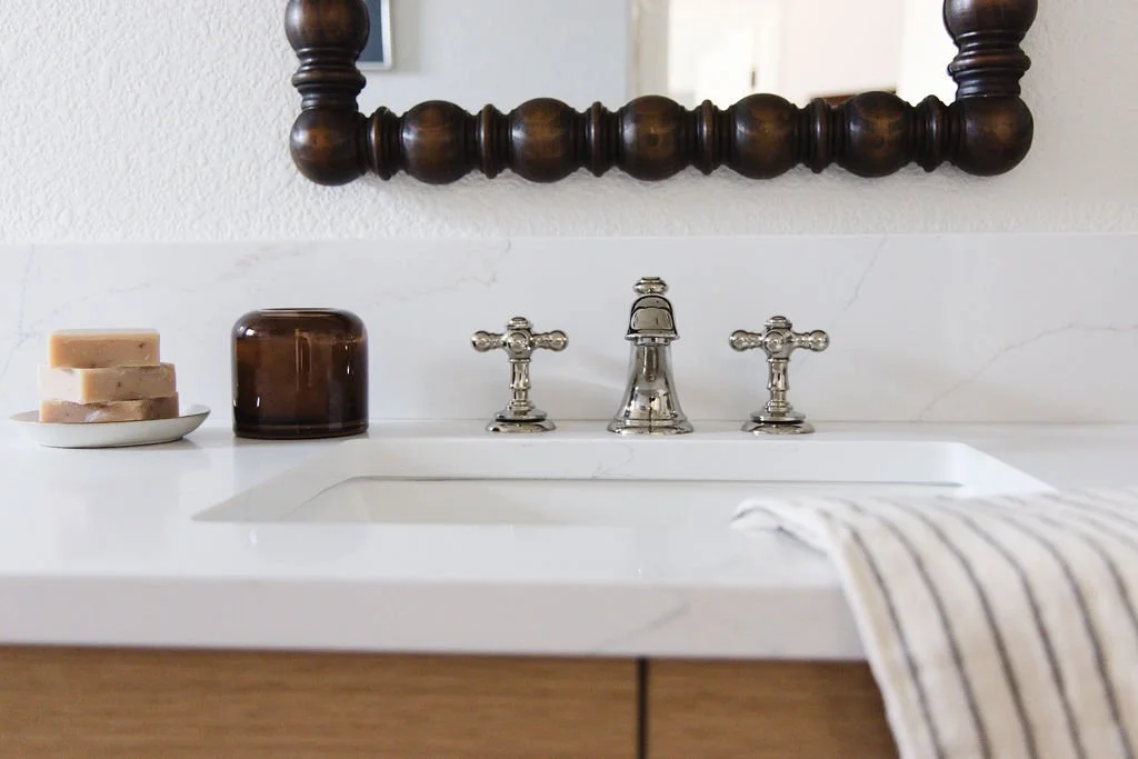 Primary bathroom with polished nickel faucet and hand-carved wood mirror, stack of hand soaps, and amber glass candle.