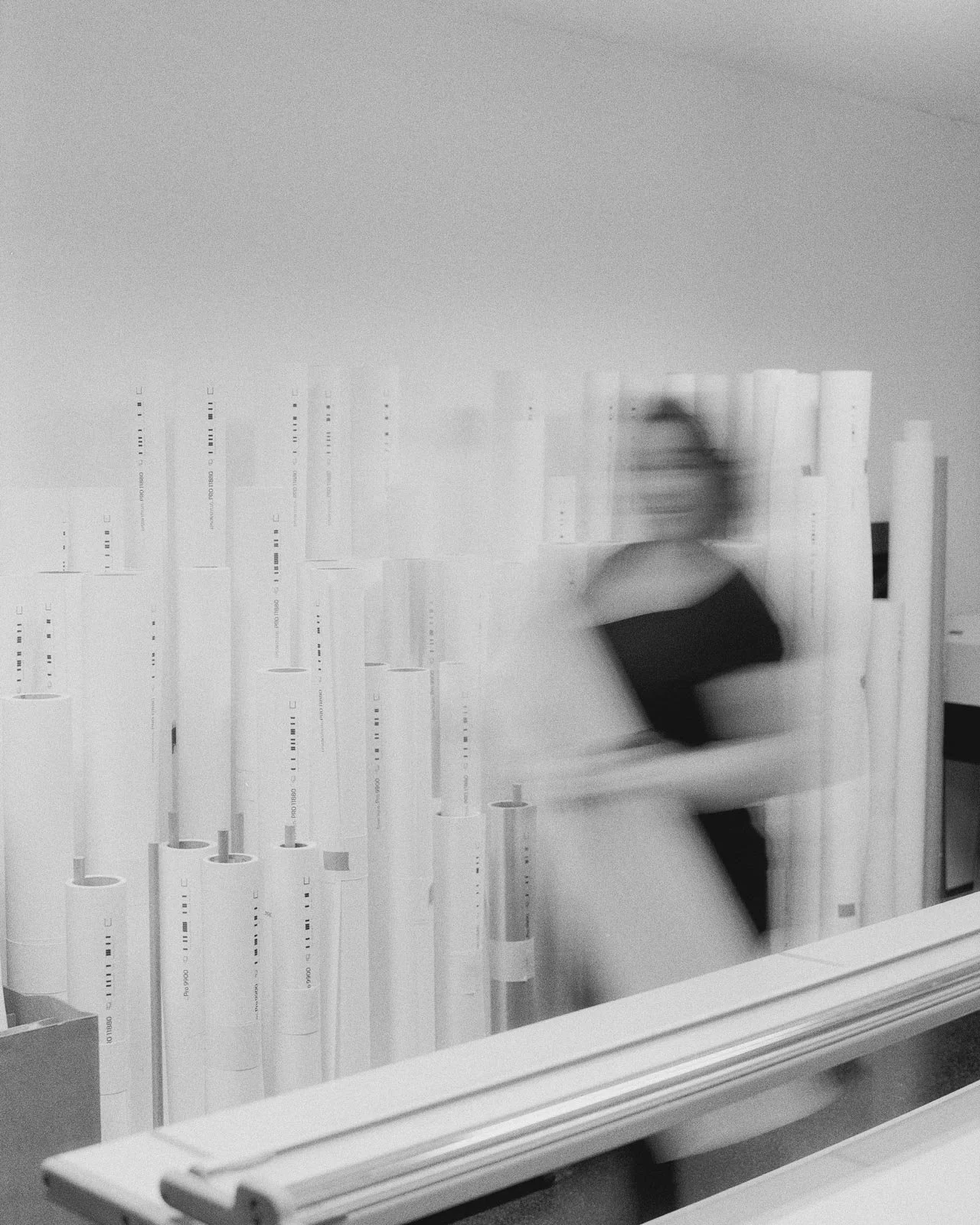Blurry black and white photo of a person sitting on a couch in front of a wall filled with rolled-up large sheets of paper or posters, with motion blur indicating movement.