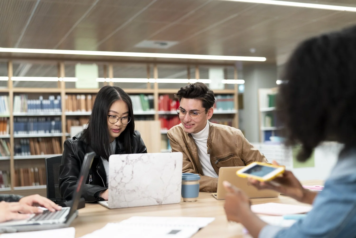 Four young adults sitting at a table in a library, working on laptops and chatting.