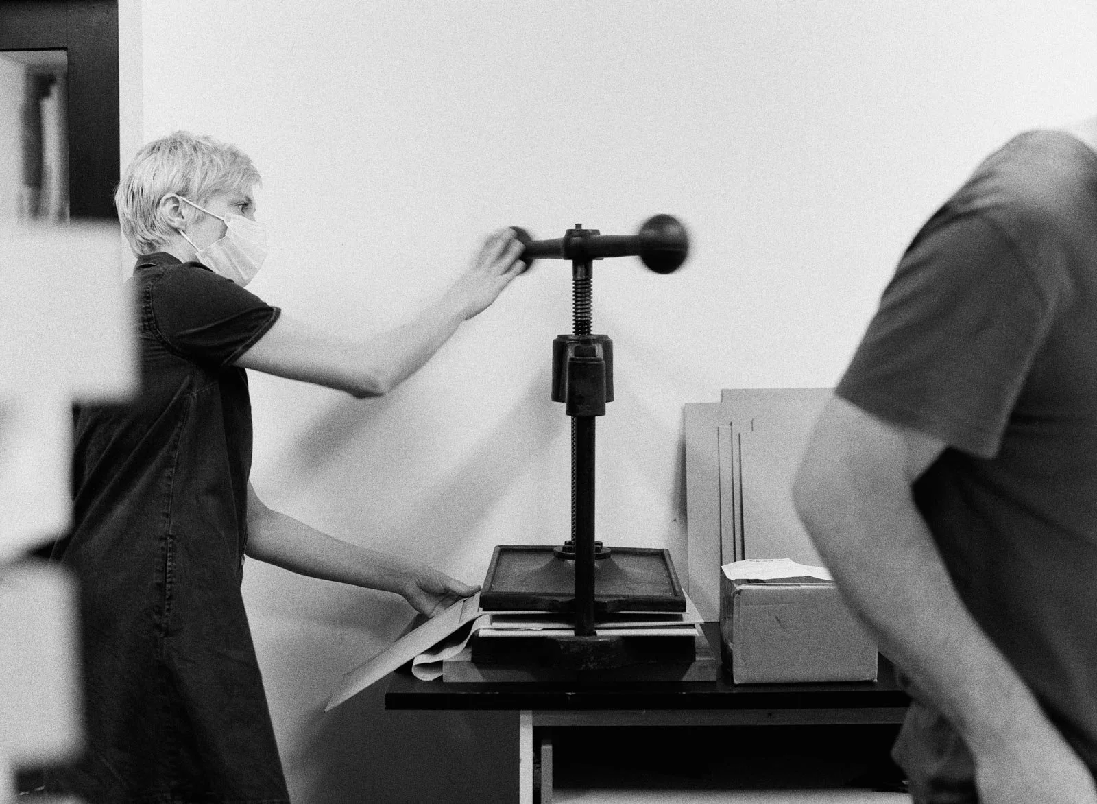 Young boy wearing a face mask operating a paper cutter or guillotine in a room with plain walls, accompanied by another person whose face is not visible.