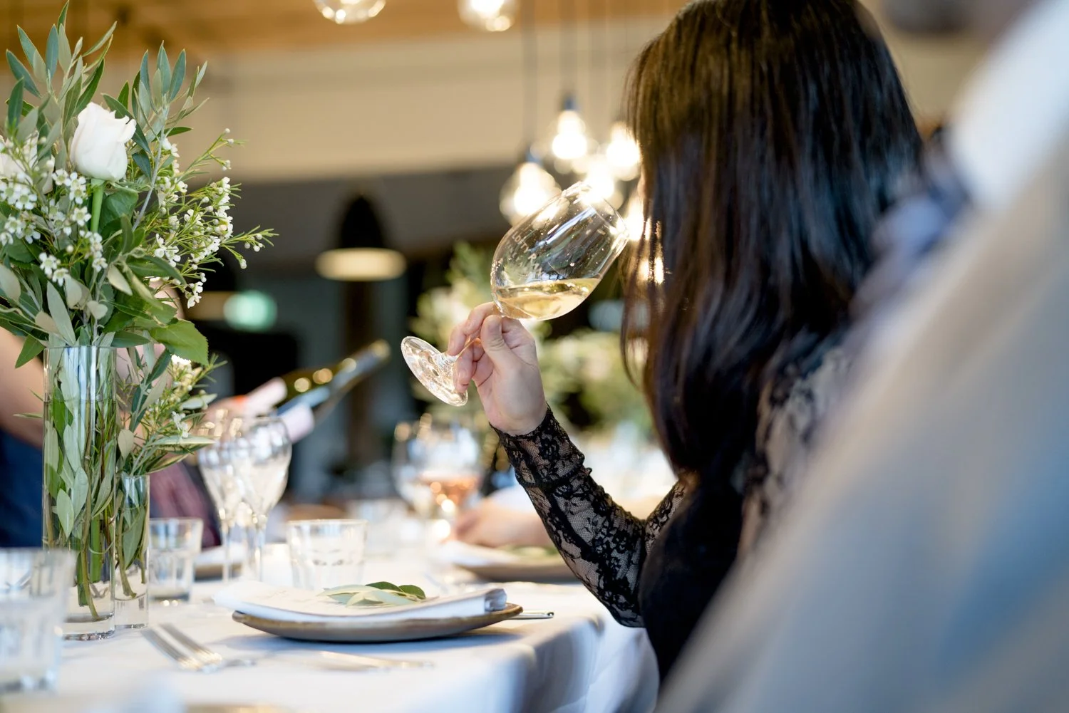Woman with dark hair and lace sleeves holding a glass of white wine at an elegant table setting with a floral centerpiece.
