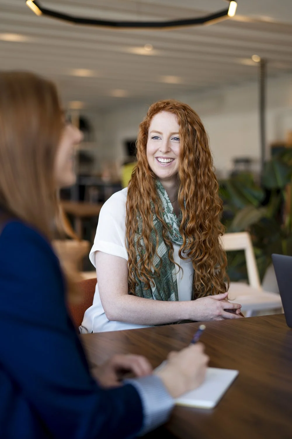 Two women having a conversation at a meeting, one with long curly red hair smiling, the other with long straight hair taking notes.