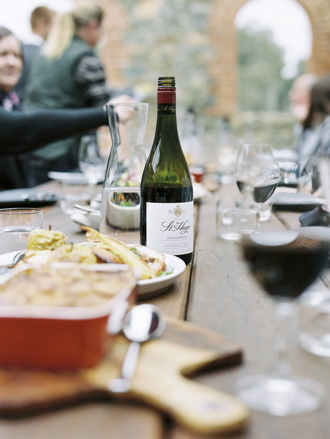 A table set with a bottle of red wine, glasses, and dishes at an outdoor gathering with people in the background.