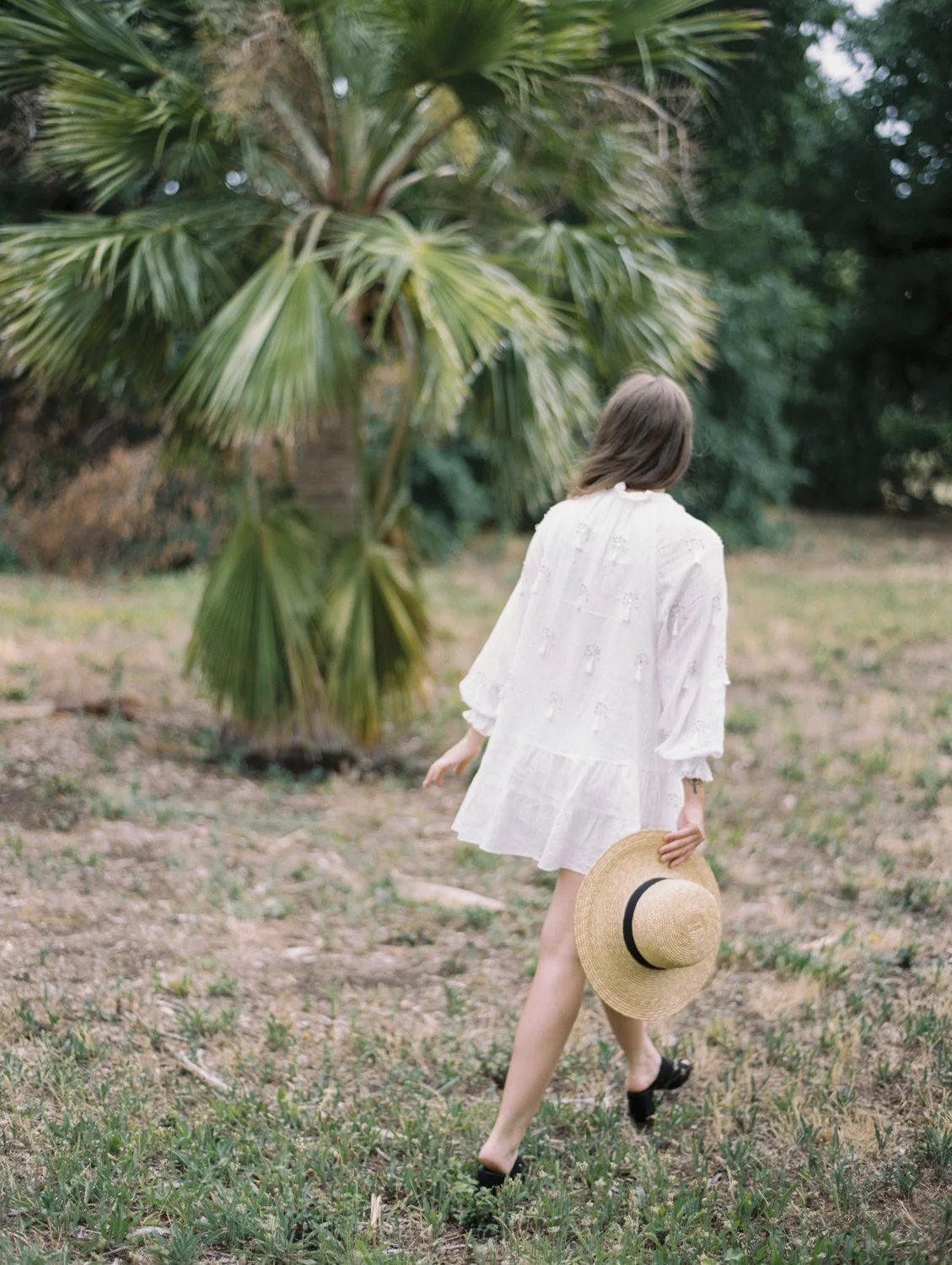 A woman in a white dress walking in a grassy area with a palm tree, holding a straw hat with a black band.