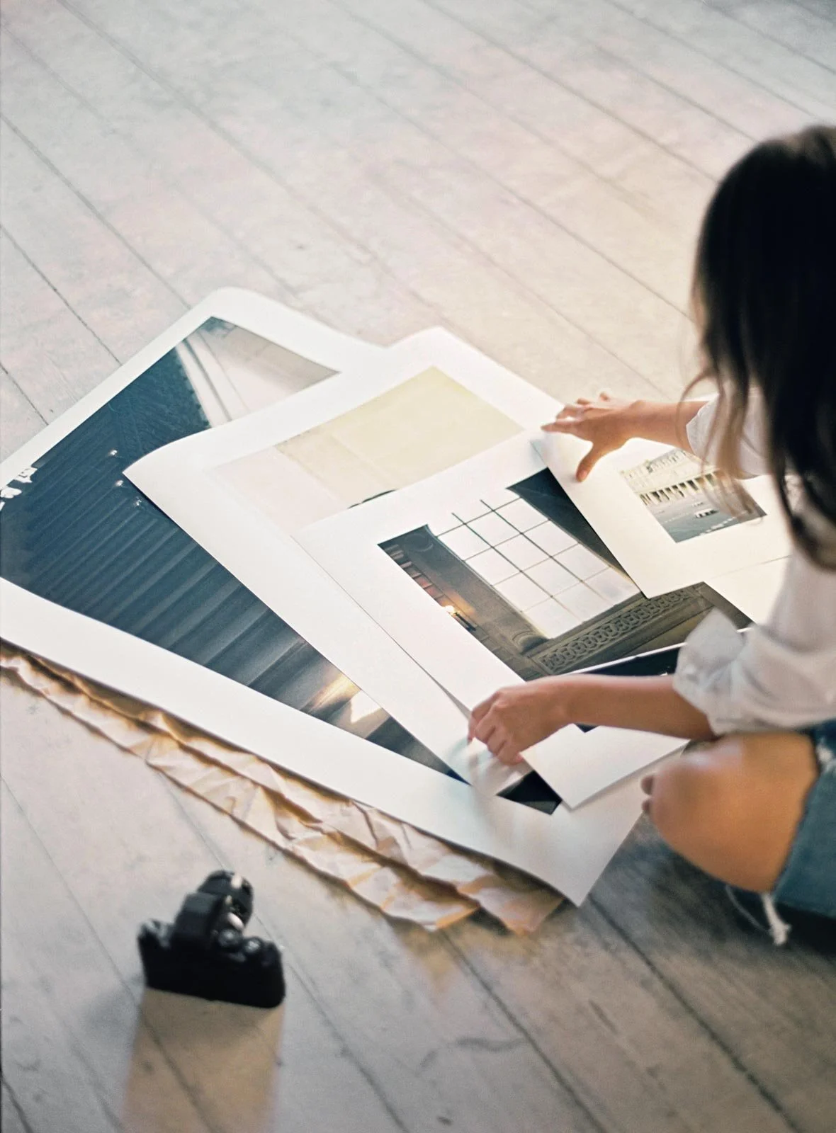 Person browsing through large photographs or artwork spread on the wooden floor, with a camera near them.