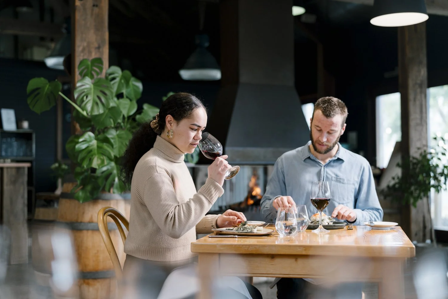 A man and woman enjoying wine at a restaurant table with a fireplace and green plant in the background.