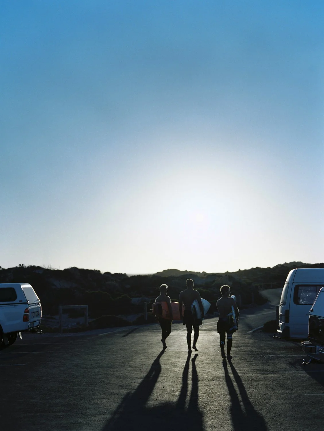 Three surfers walking towards the ocean at sunset with surfboards, cars parked nearby, and a clear sky.