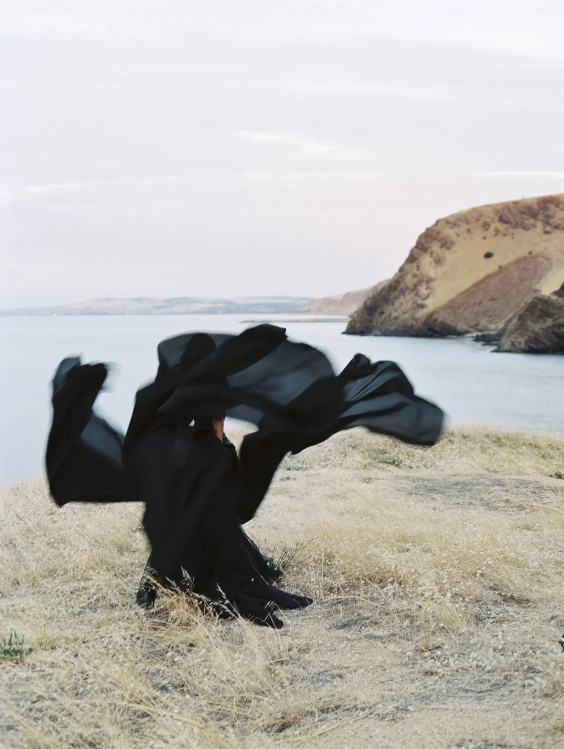 A person dressed in black is sitting on dry grass near a body of water with hills or cliffs in the background, holding up a flowing piece of black fabric.