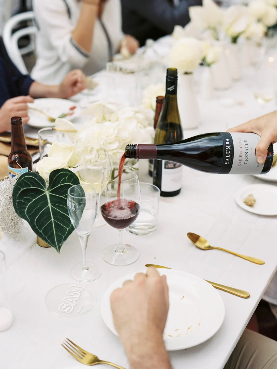 Person pouring red wine into a glass on a decorated dining table with white roses and gold cutlery.