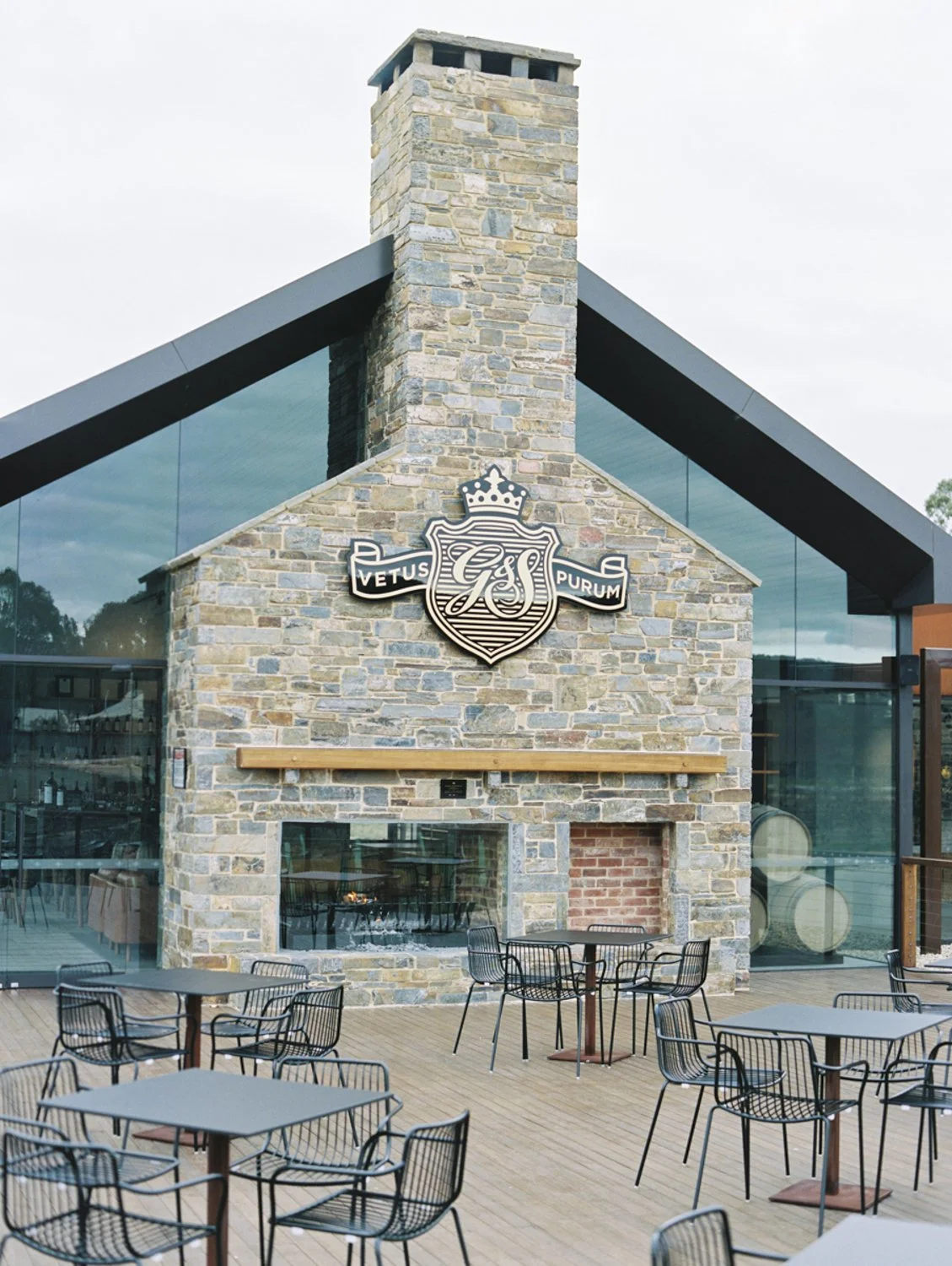 Outdoor patio with tables and chairs in front of a building with a stone chimney and glass walls. The building displays a crest with the Latin words "Vetus Purum."