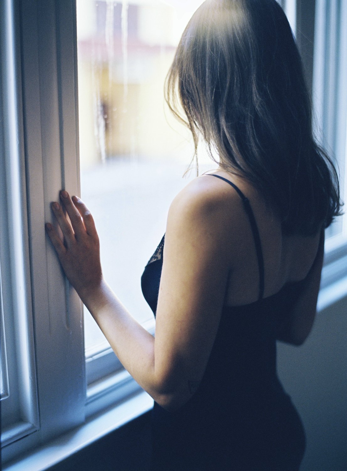 A woman with shoulder-length brown hair, wearing a black spaghetti strap top, stands by a window with her hand on the window frame, gazing outside.