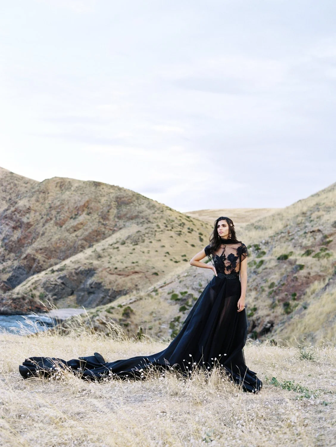 A woman in a black dress with a long train stands in a dry, grassy field with rolling hills and a river in the background.
