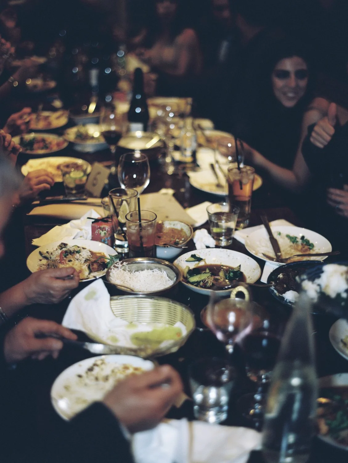 A table full of partially eaten food, drinks, and plates at a social gathering, with people around it, in a dimly lit setting.
