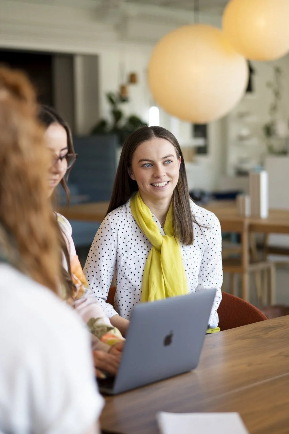 Three women gathered around a wooden table in a modern, well-lit room. One woman with long brown hair and a yellow scarf is smiling and looking to her left. Another woman with brown hair and glasses is partially visible, using a silver laptop. The th