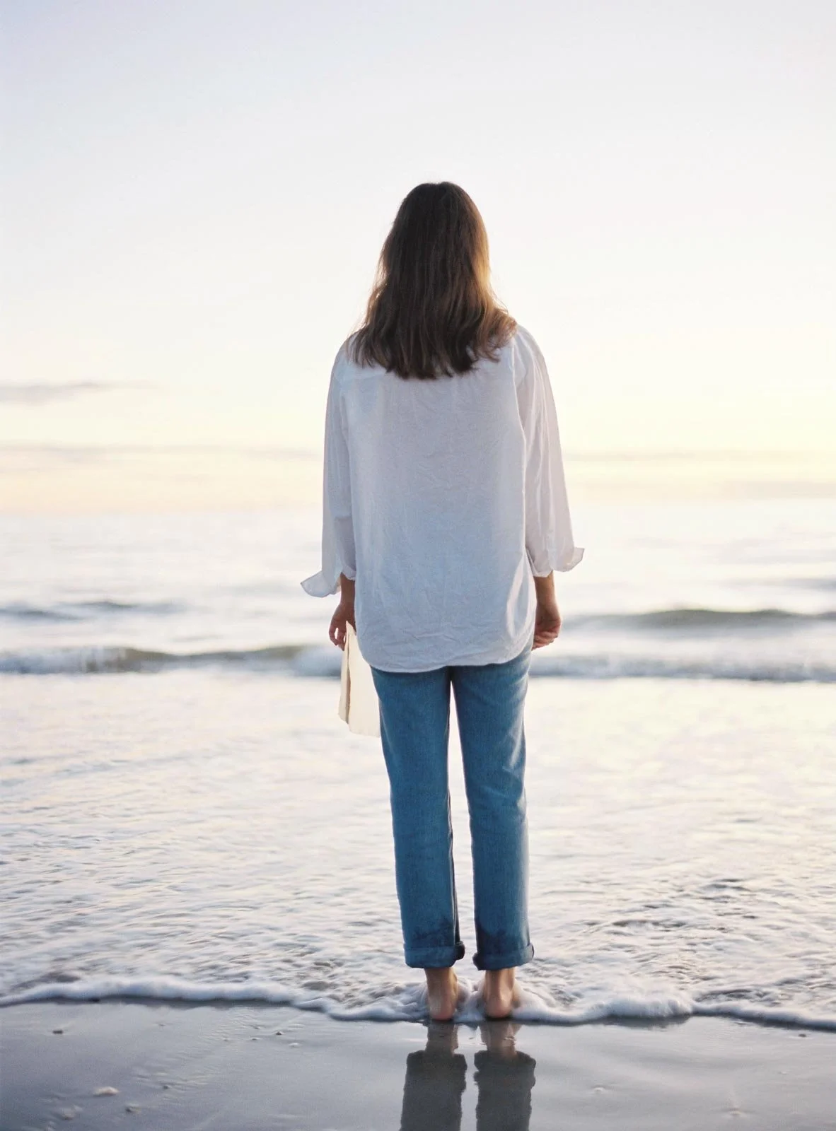Woman standing barefoot at the edge of the ocean, facing the water during sunset or sunrise.